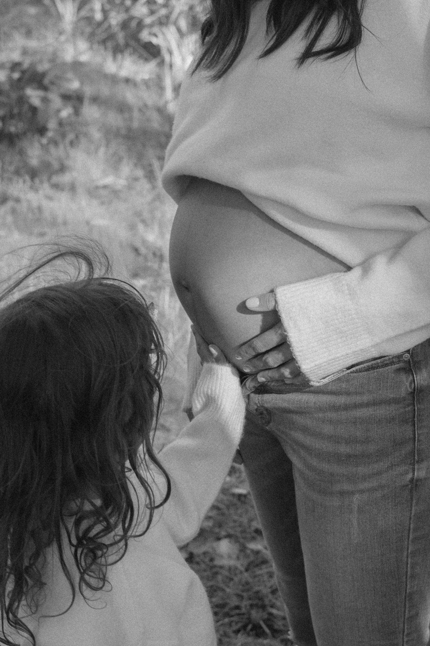 Black and white photo of pregnant woman with child touching her bare baby bump outdoors.