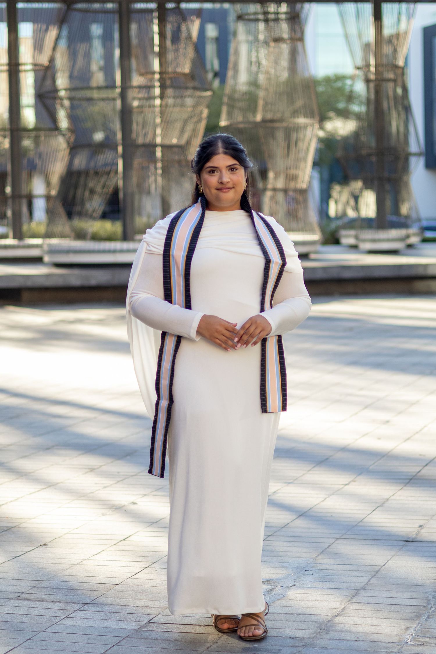 A young Indian woman in a white traditional dress with a striped dupatta posing outdoors.