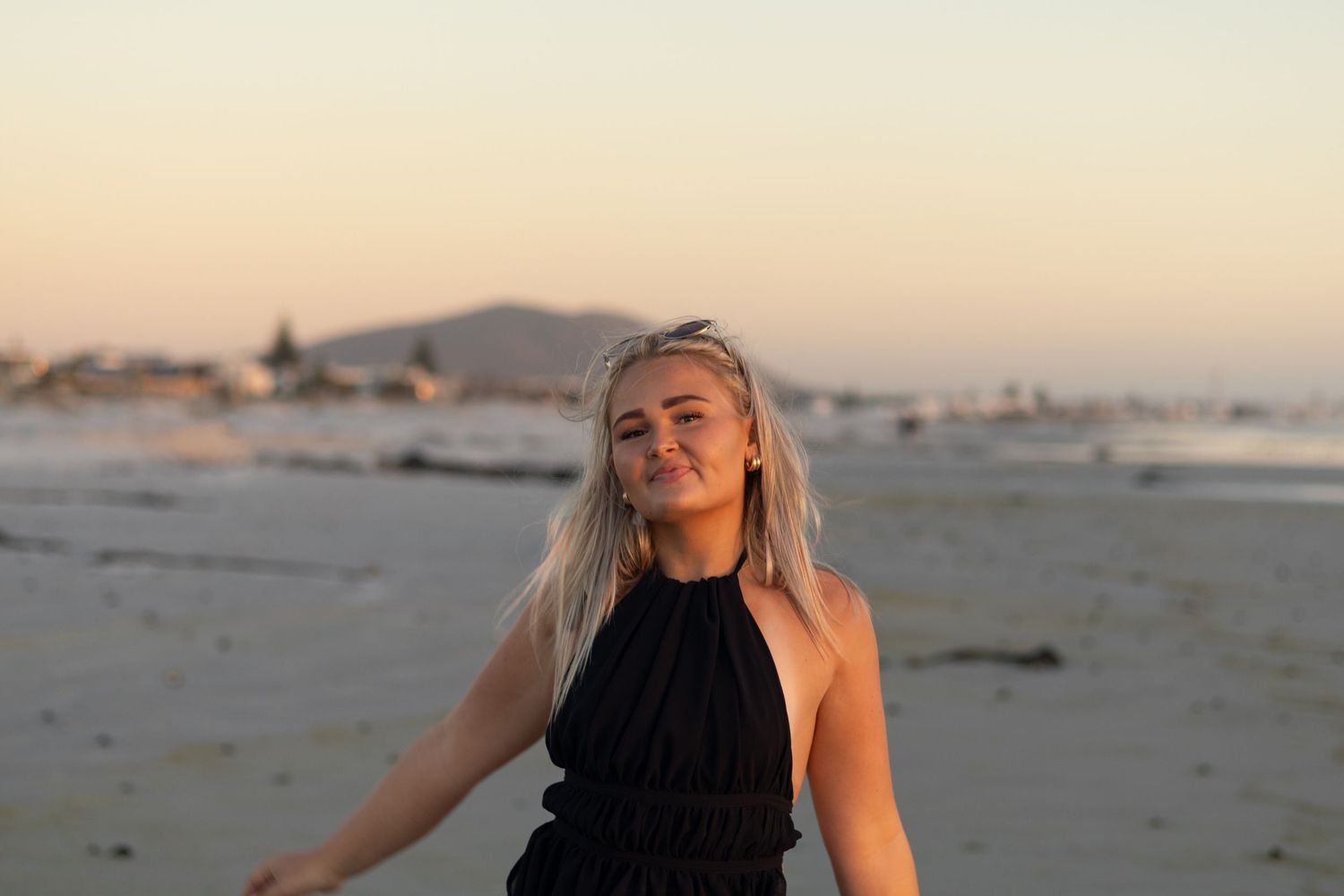 Young blonde woman in black halter dress posing on a sandy beach at golden hour sunset.