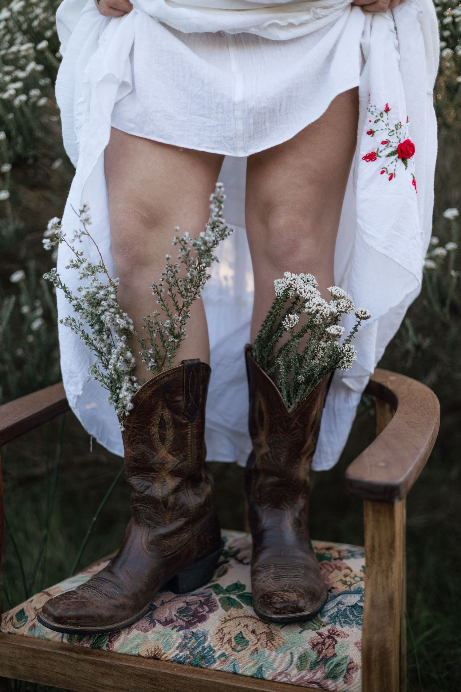 Person in white dress and cowboy boots with wildflowers, standing on a vintage chair in a flower field.