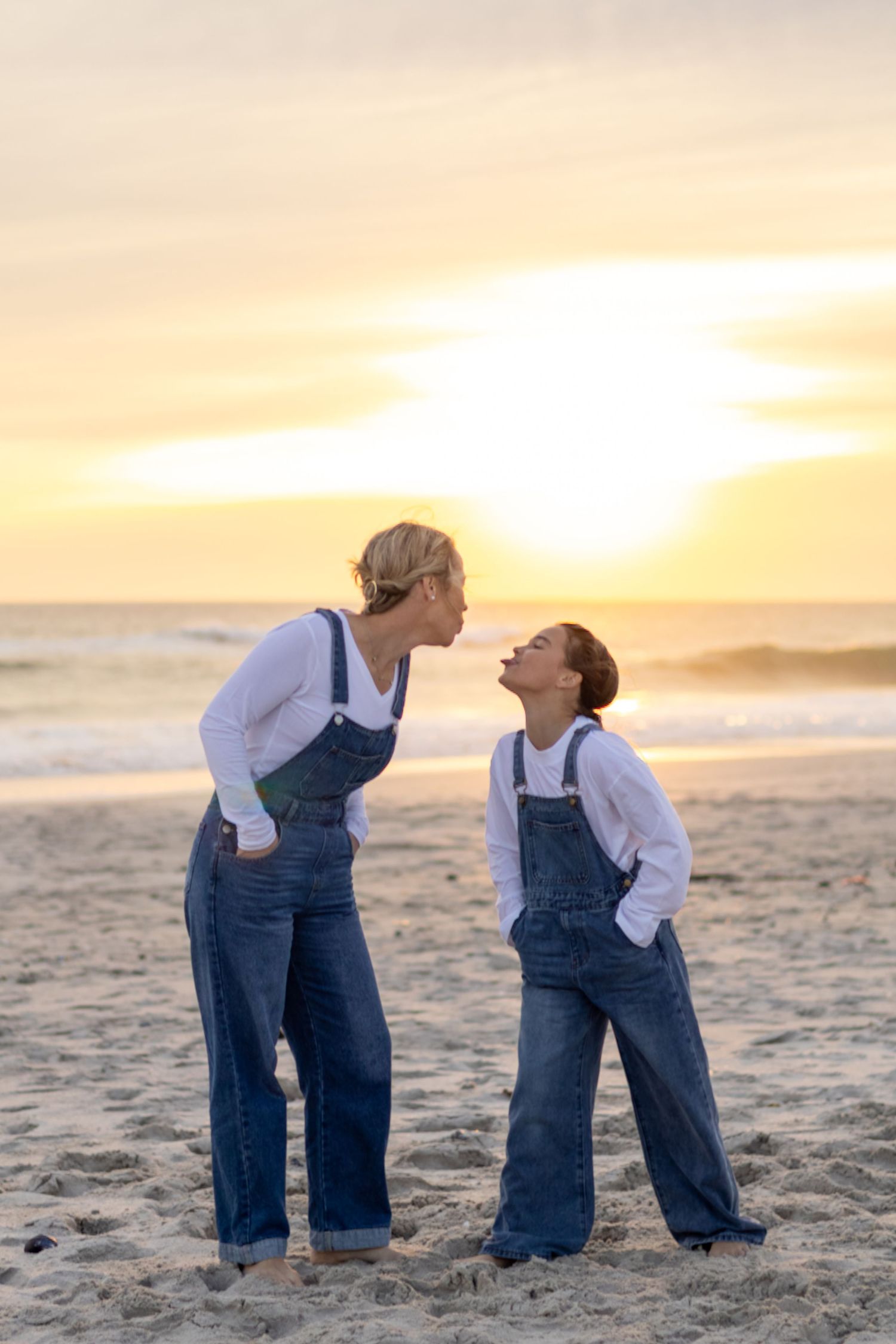 Mother and daughter in matching denim overalls laughing together on a sandy beach at golden sunset.