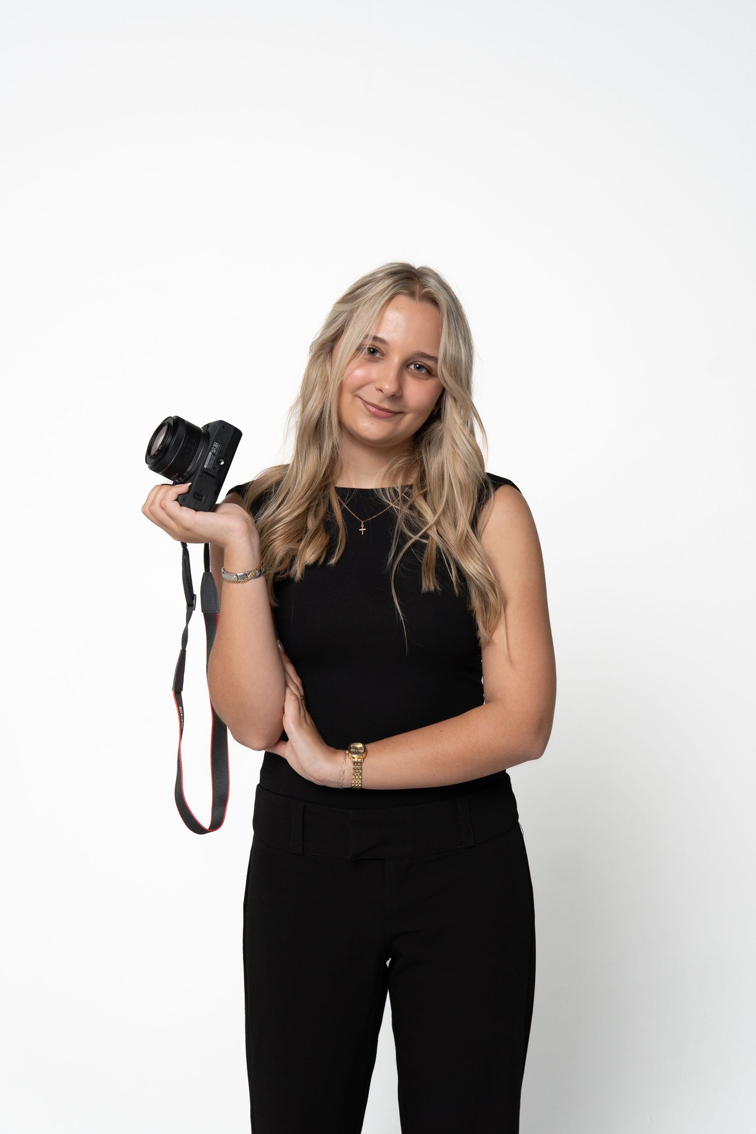 Young blonde woman in black outfit holding a DSLR camera, smiling against a white background.