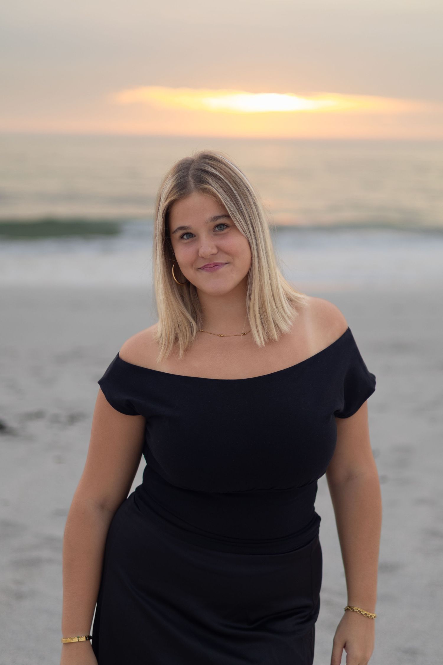A blonde woman in a black off-shoulder dress posing on a sandy beach at golden sunset.
