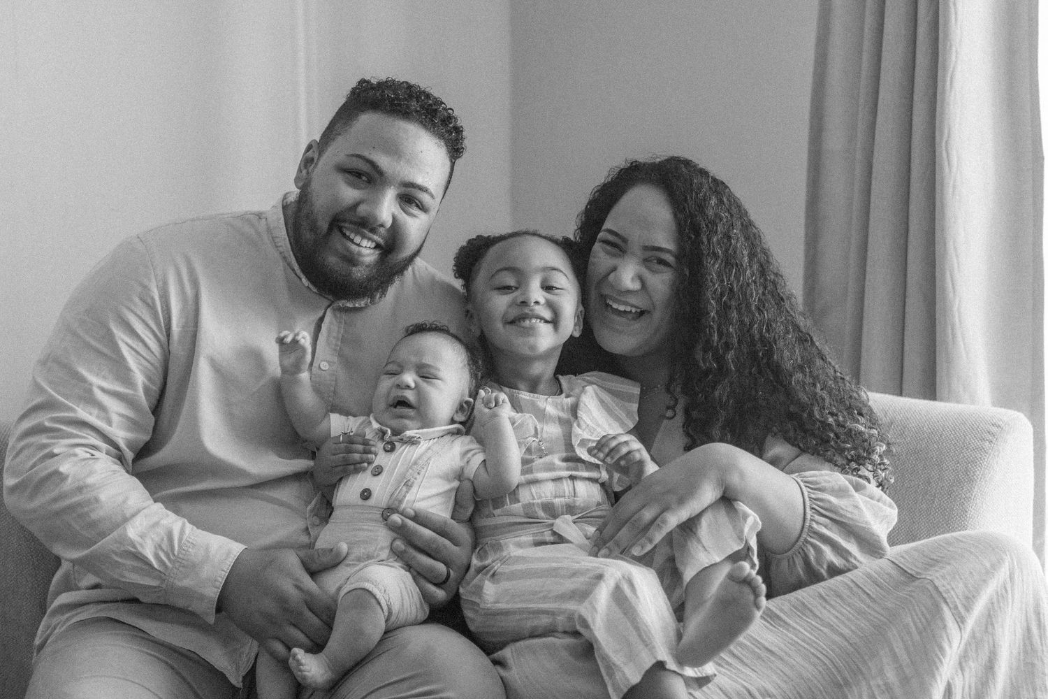 Black and white family portrait of parents smiling with toddler and baby sitting together on a couch.