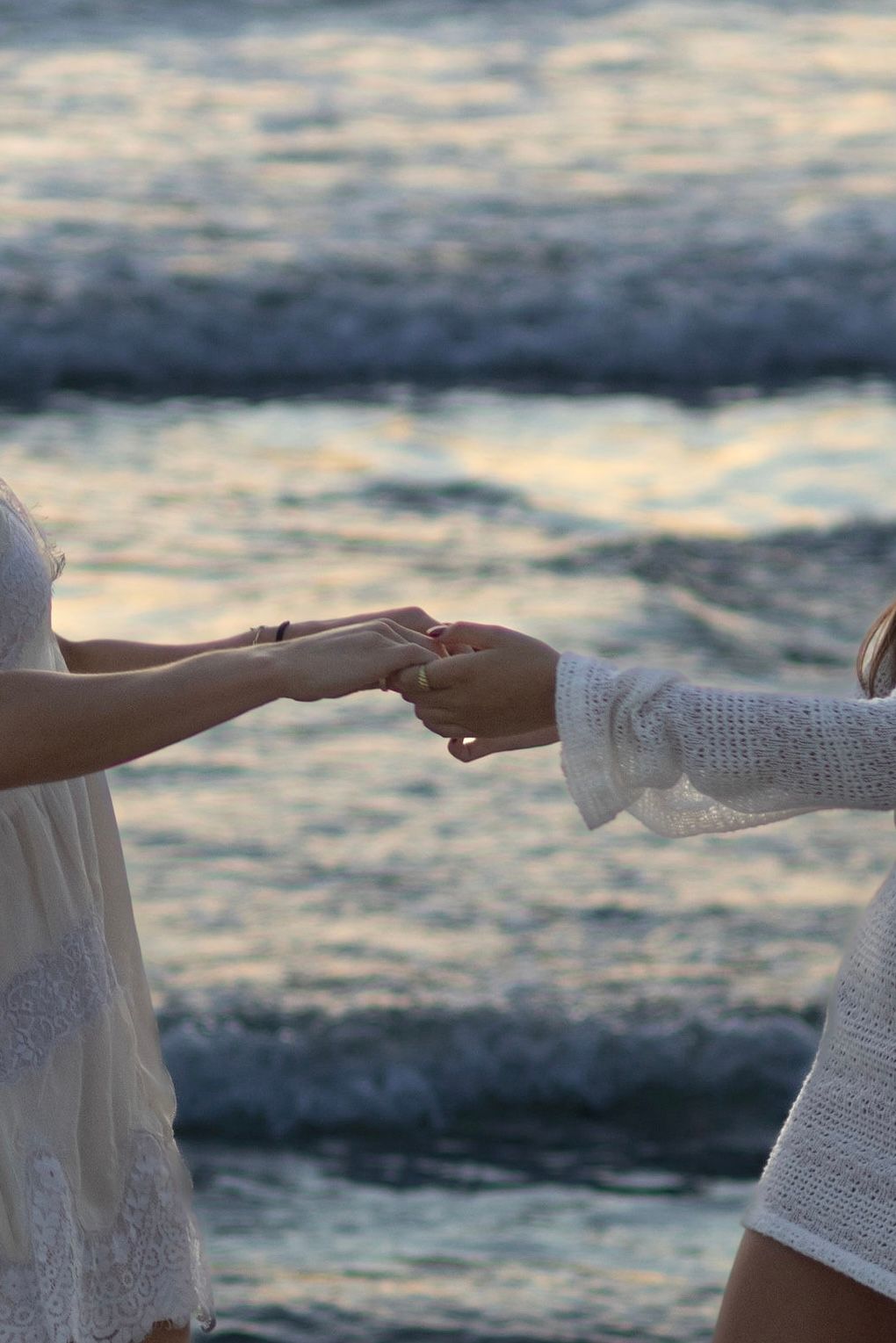 Two women in white dresses holding hands near ocean waves at sunset.