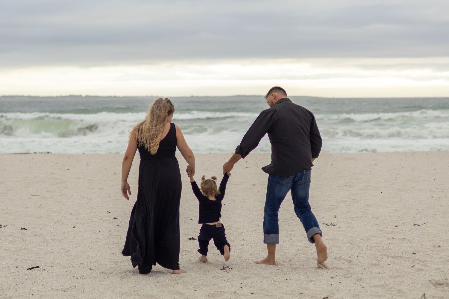 Family of three holding hands walking on a sandy beach with ocean waves and cloudy sky in the background.