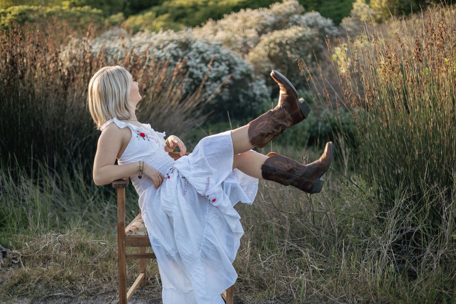 Woman in white dress and cowboy boots sitting on wooden chair in field, kicking leg up playfully.
