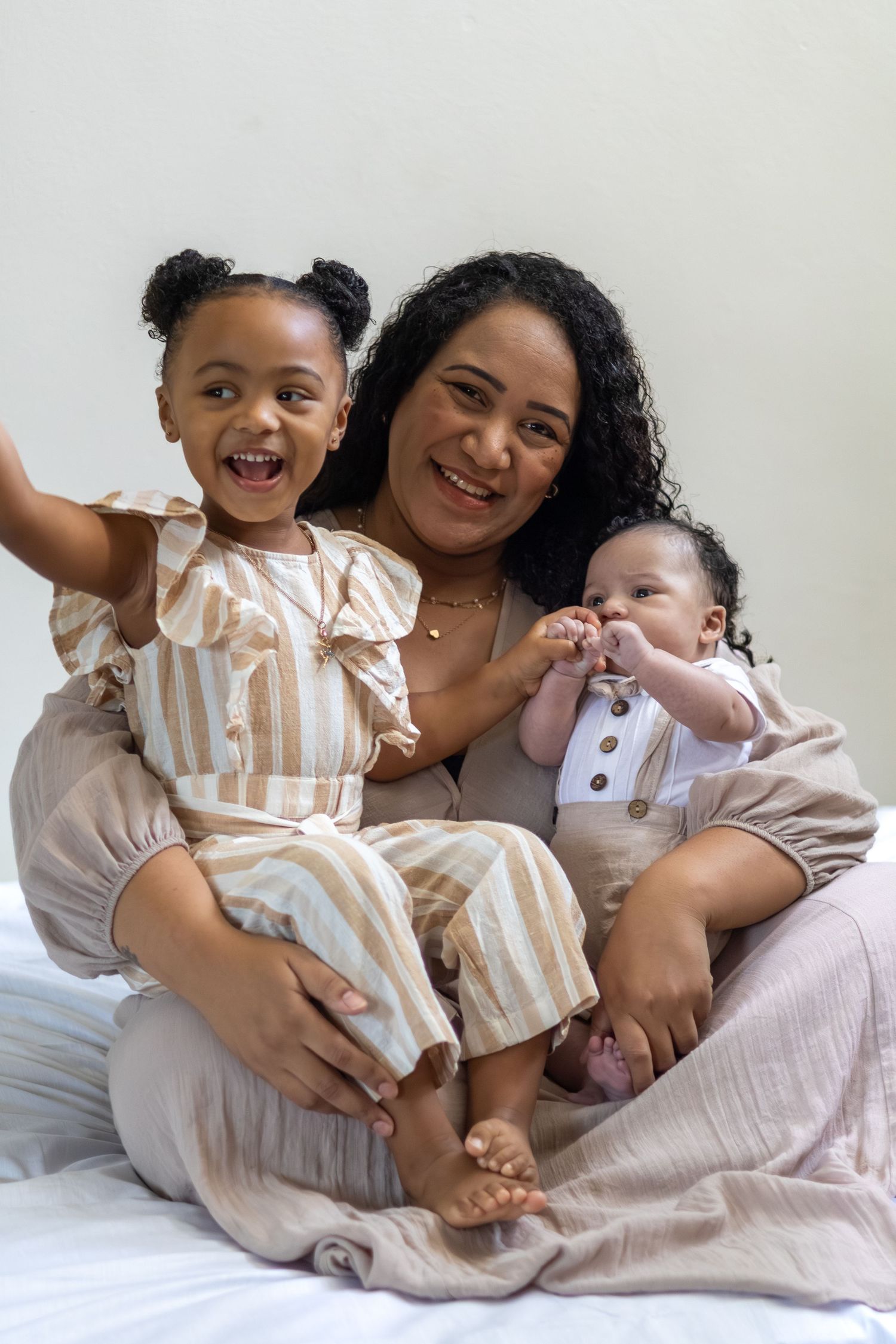 A smiling mother sits on a bed holding her toddler daughter and baby son, all dressed in matching neutral tones.