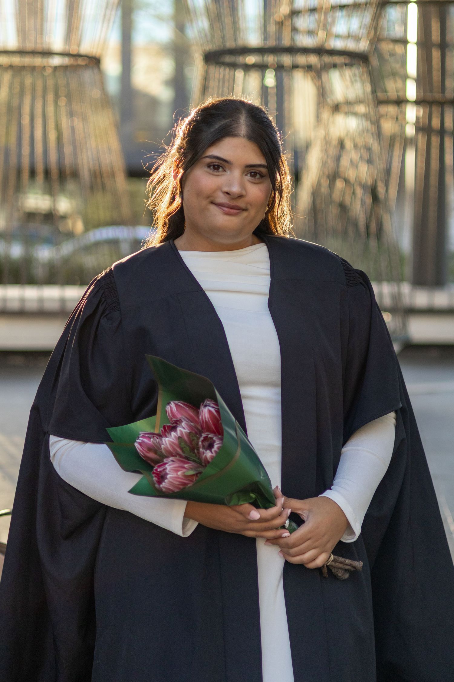 A smiling graduate in a black gown holds pink tulips, posing outdoors in a white dress at her graduation ceremony.