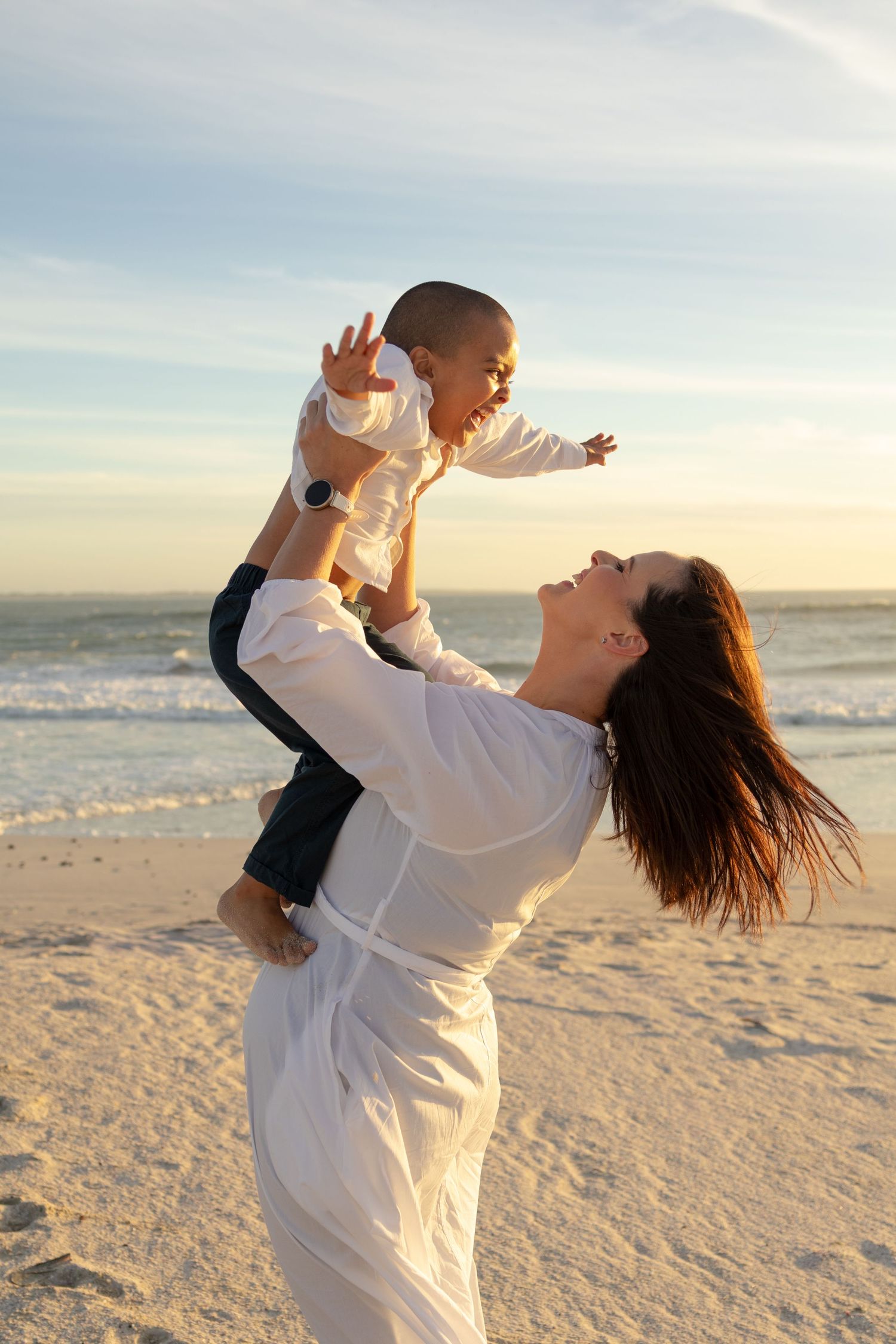 A joyful mother lifts her baby high at a sunny beach, both smiling with ocean waves in the background.