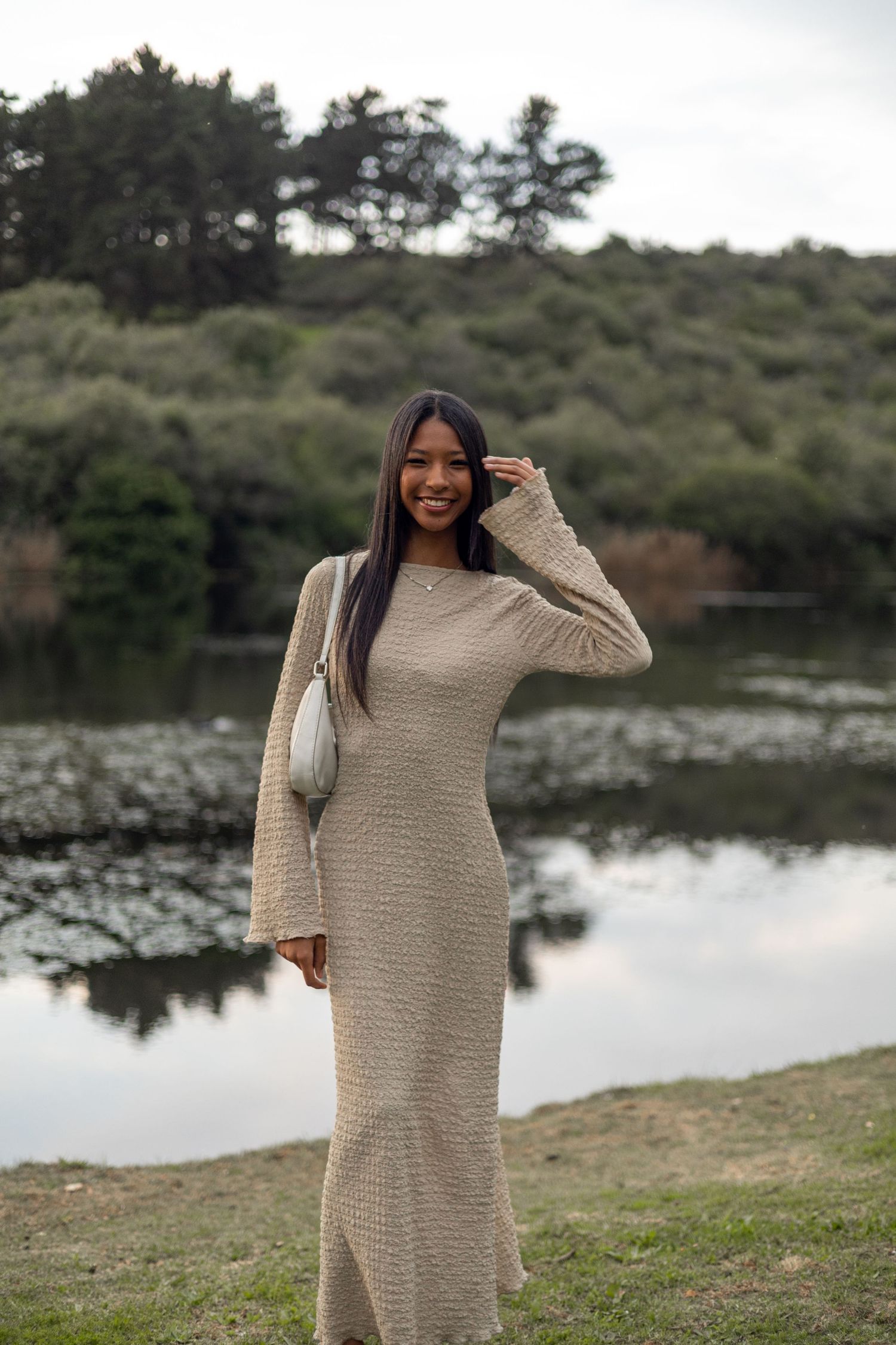 Woman in beige knit maxi dress posing by a reflective lake with lush green trees in the background.
