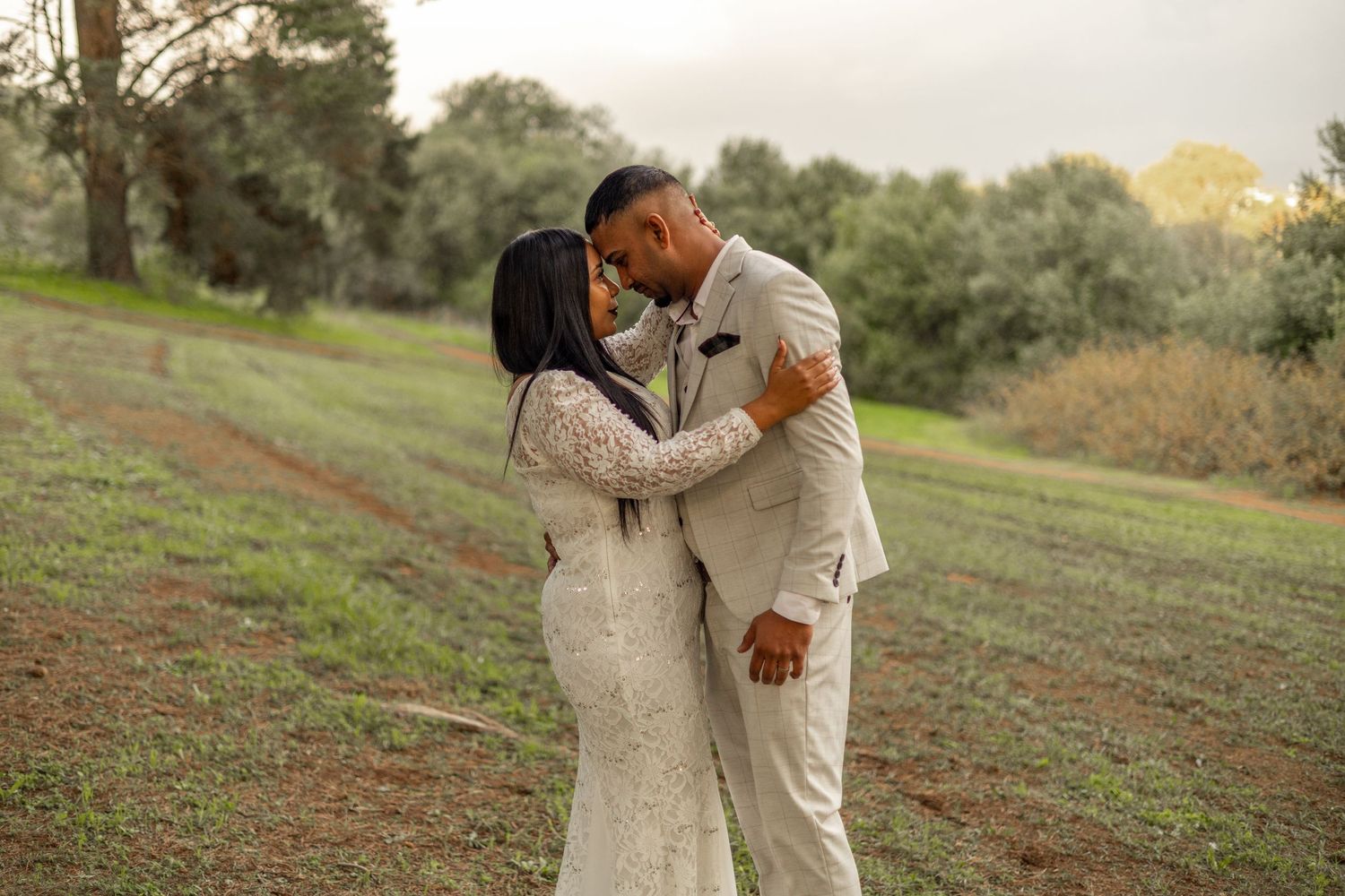 A couple shares a romantic kiss in a lush green field, bride in lace gown and groom in beige suit.