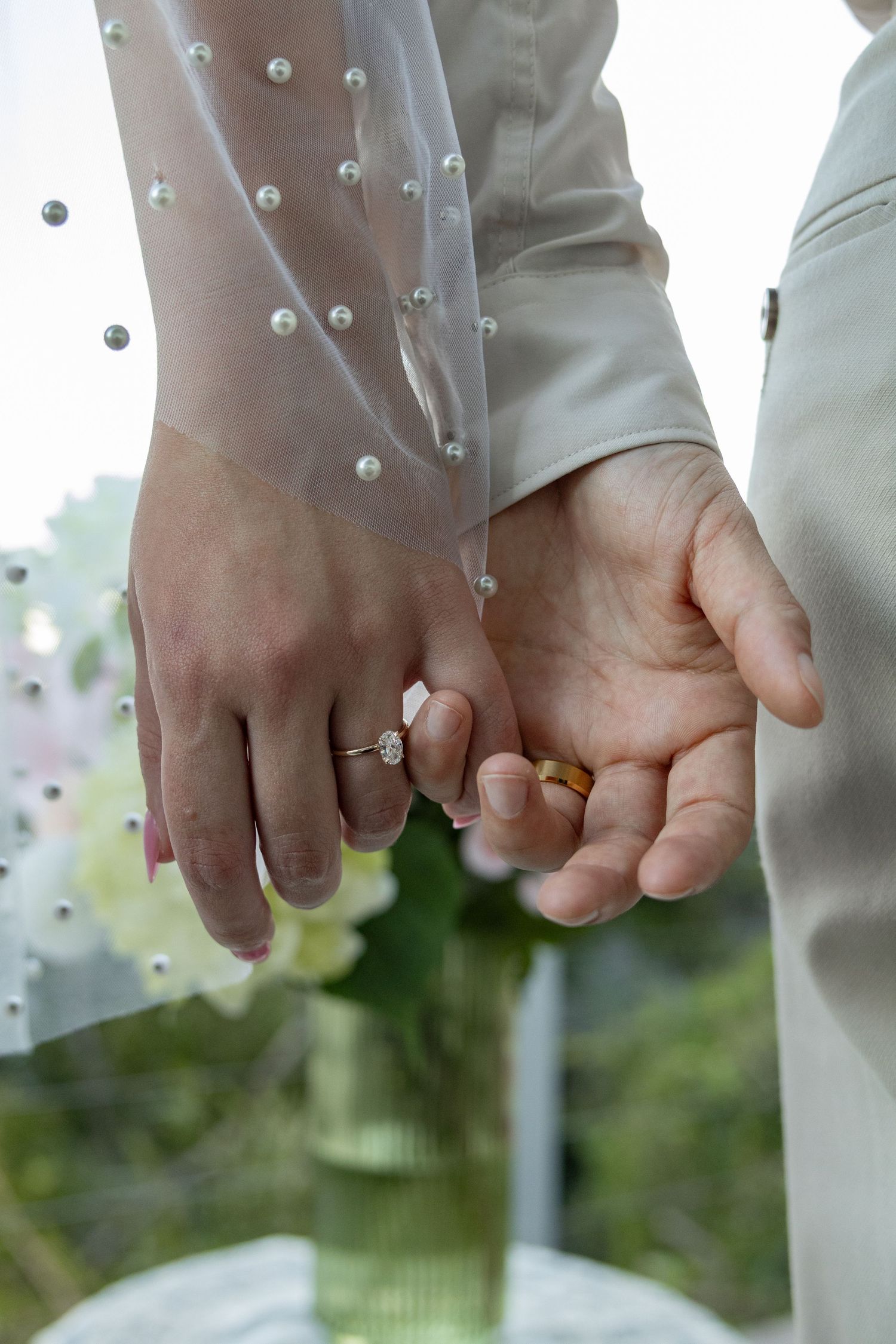 Bride and groom holding hands, showcasing diamond engagement ring and gold wedding band at outdoor ceremony.