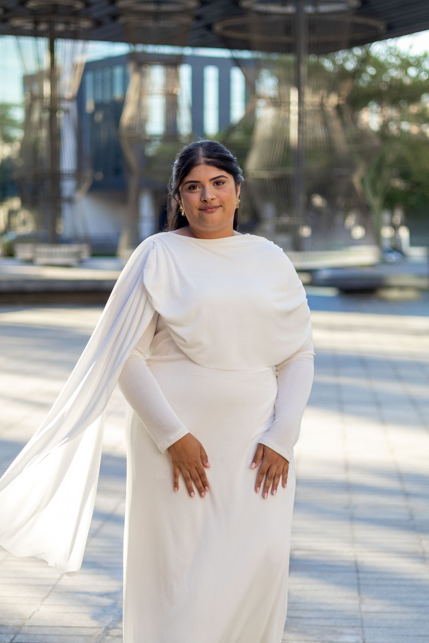 Woman in elegant white long-sleeve gown with flowing cape poses outdoors in urban setting.