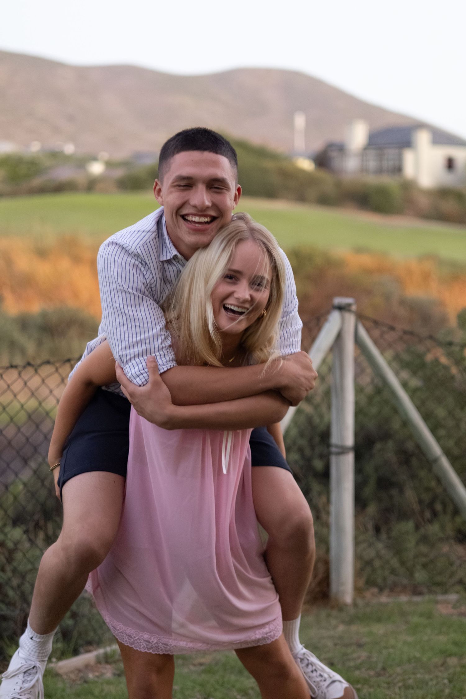 A laughing couple enjoying a piggyback ride outdoors with green hills in the background.