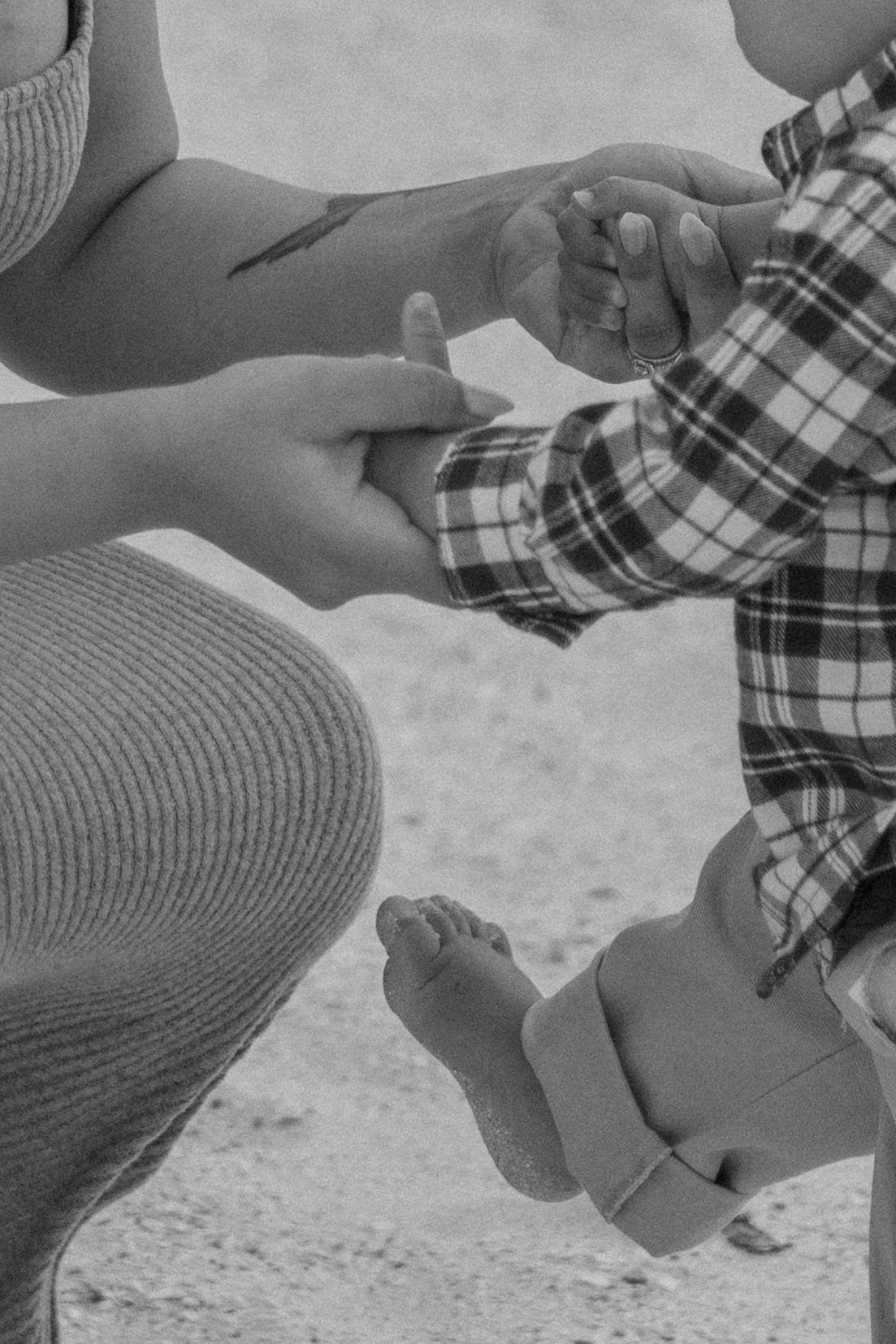 Black and white photo of a mother holding her baby's hands on a sandy beach.