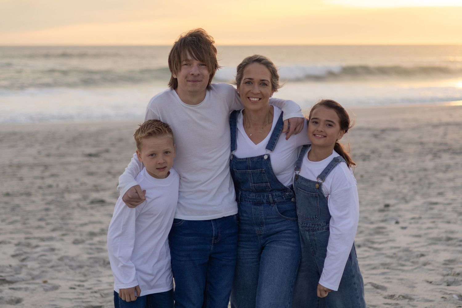 Family of four smiling together on a sandy beach at sunset, wearing white tops and denim outfits.