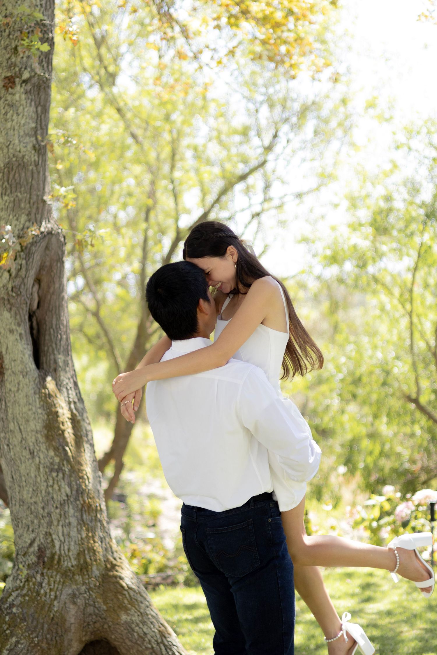Romantic couple embracing in a sunny park, man lifting woman in white dress among green trees.