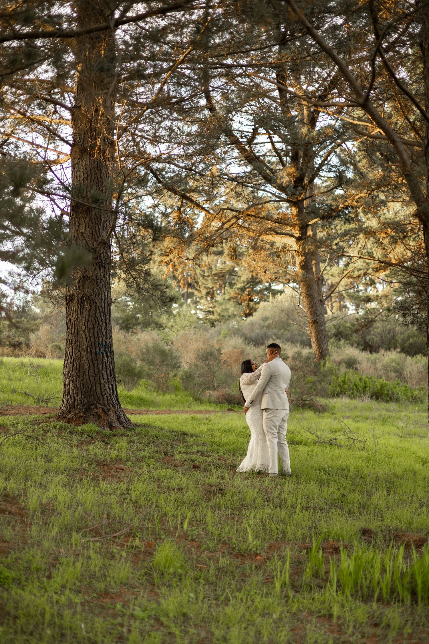 Couple in white attire embracing under tall pine trees in a lush green meadow during golden hour.