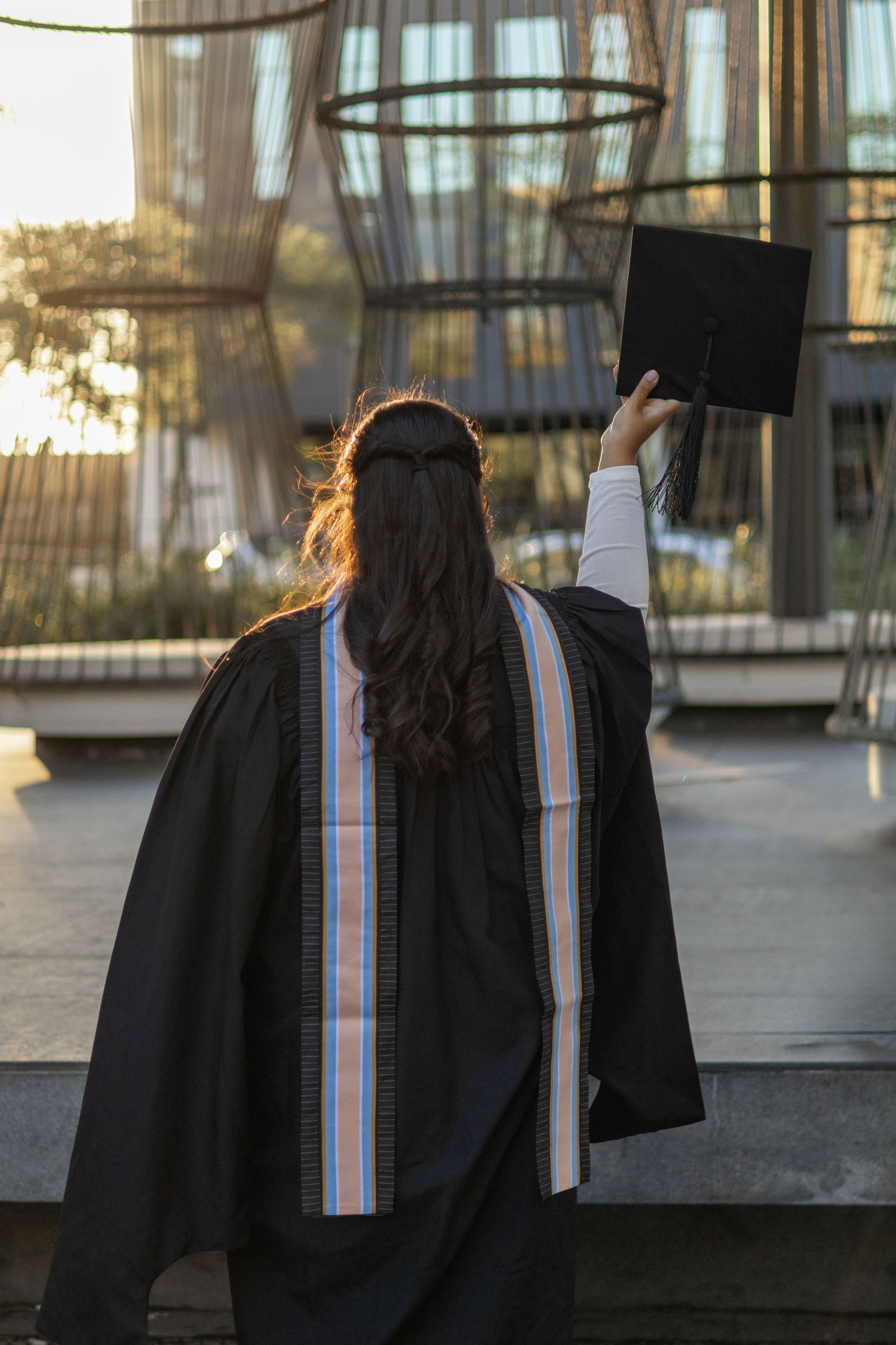 Graduate in black gown holding cap aloft, seen from behind, with long hair and colorful stole outdoors.