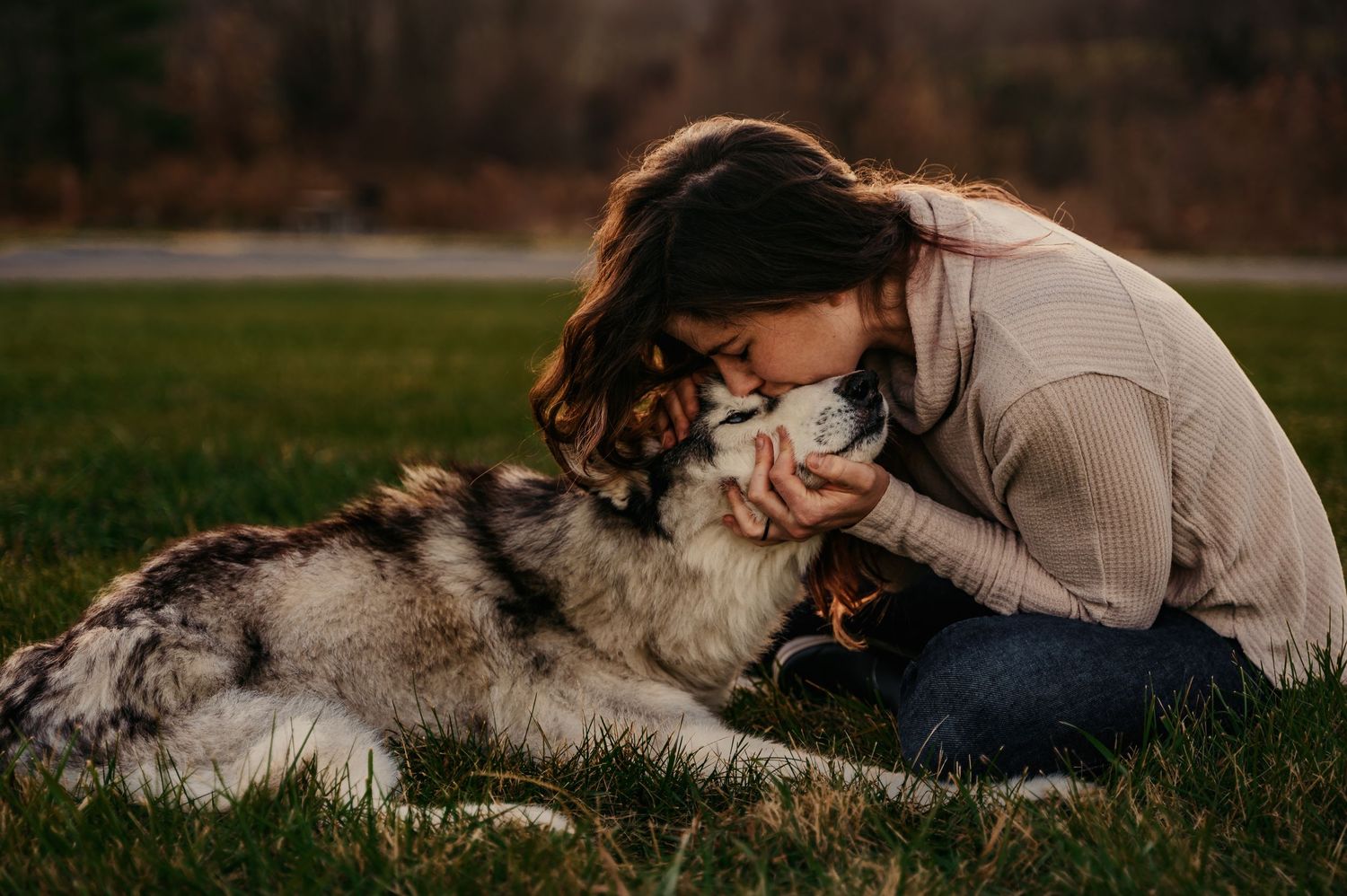 Young woman sitting on grass, lovingly nuzzling her fluffy Husky dog outdoors in a park setting.
