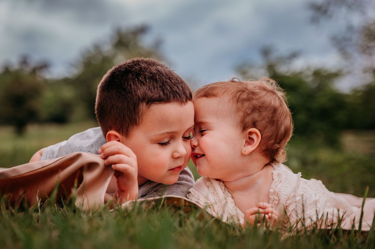 Two young siblings lying in green grass, touching foreheads and sharing a sweet tender moment outdoors in Winchester Virginia