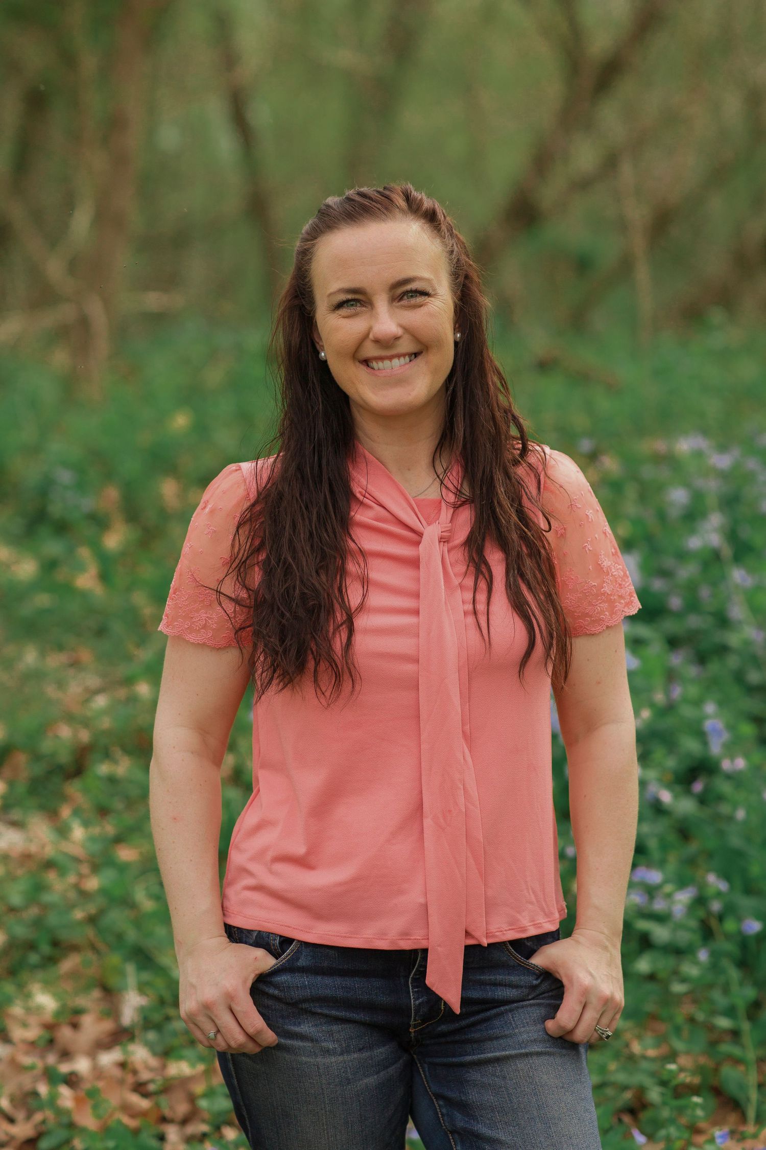 Smiling woman with long dark hair wearing a coral pink blouse standing outdoors in a lush green woodland setting.