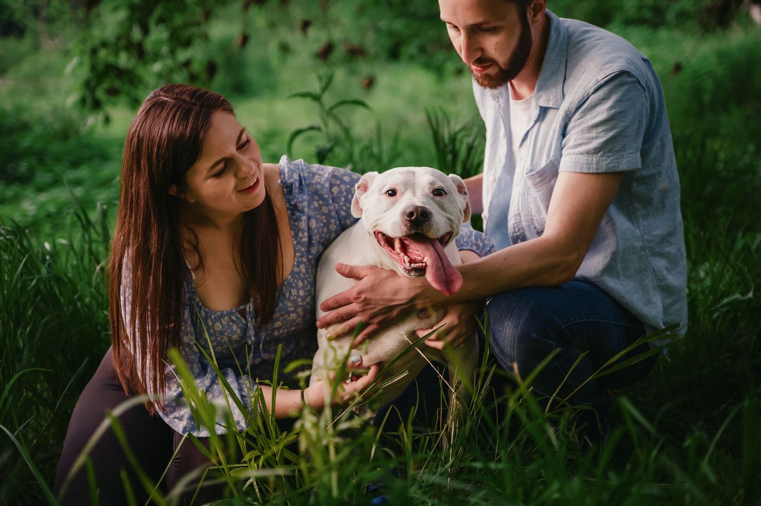 A couple sits in lush green grass with their happy white dog, enjoying a relaxed outdoor moment together on the Shenandoah River