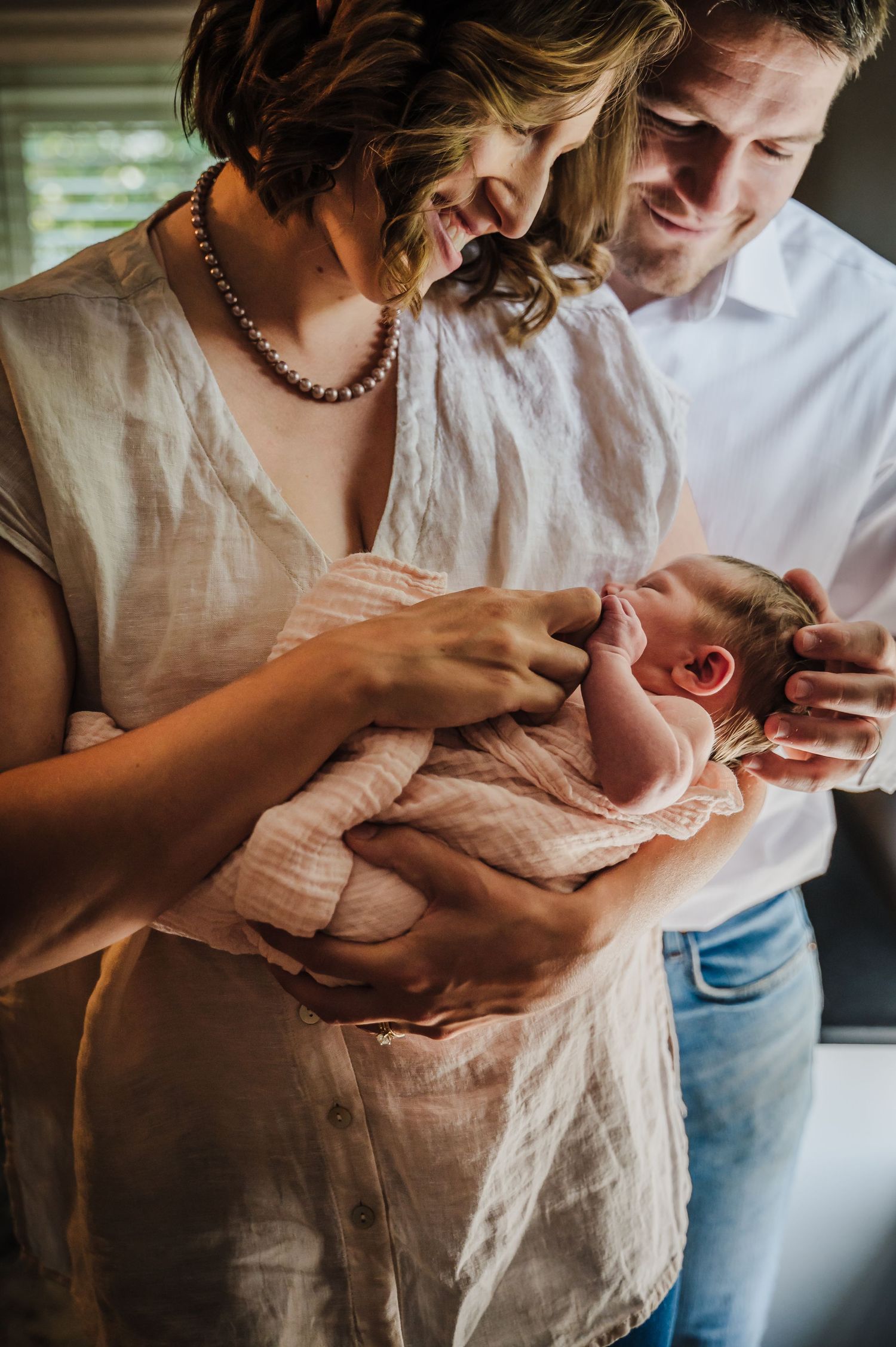 New parents lovingly hold their newborn baby wrapped in a pink blanket, sharing a tender family moment indoors.