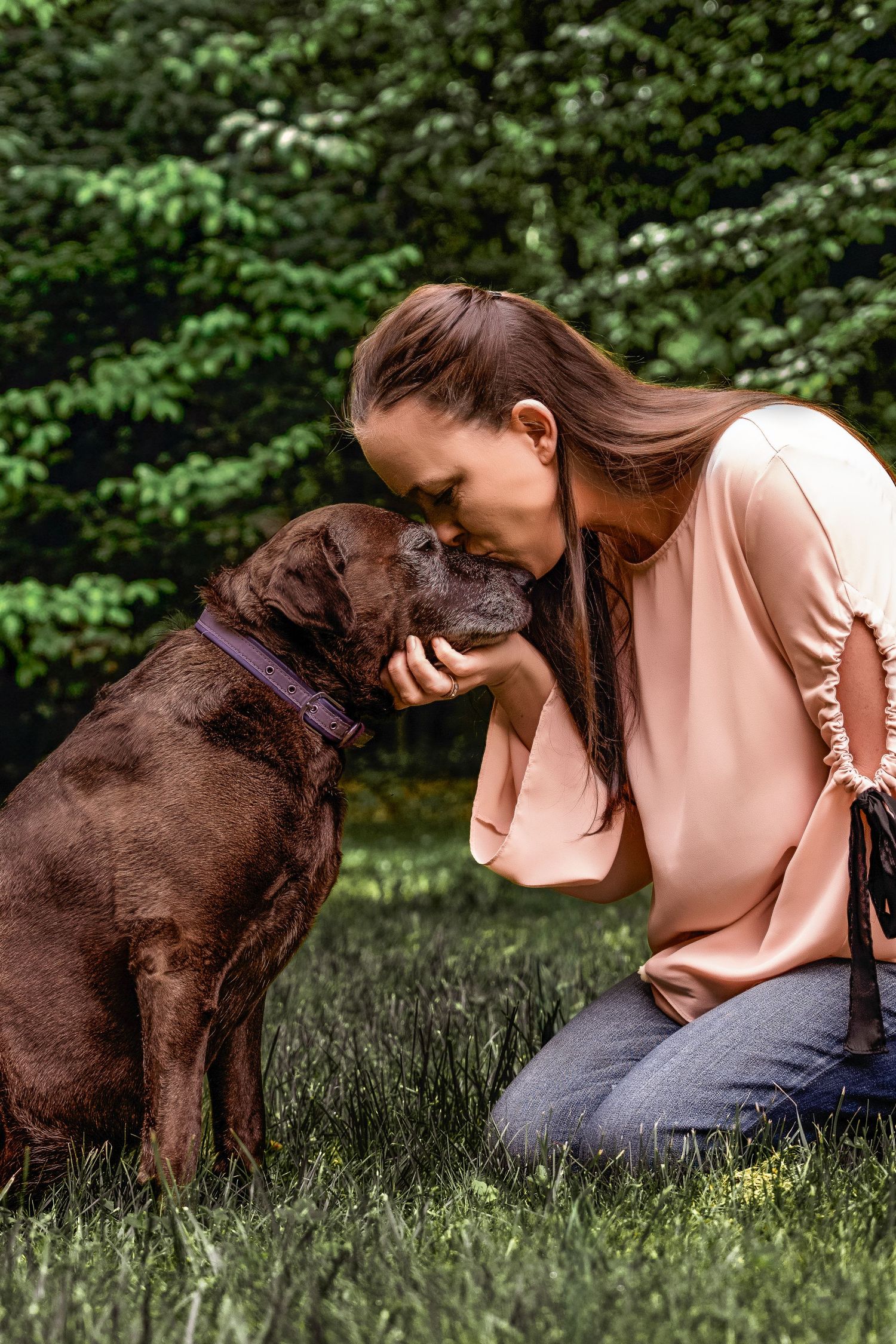 Photographer Kayleigh Coffman kneeling in the grass and kissing her senior dog Kenzie on the forehead in a wooded park in Northern Virginia