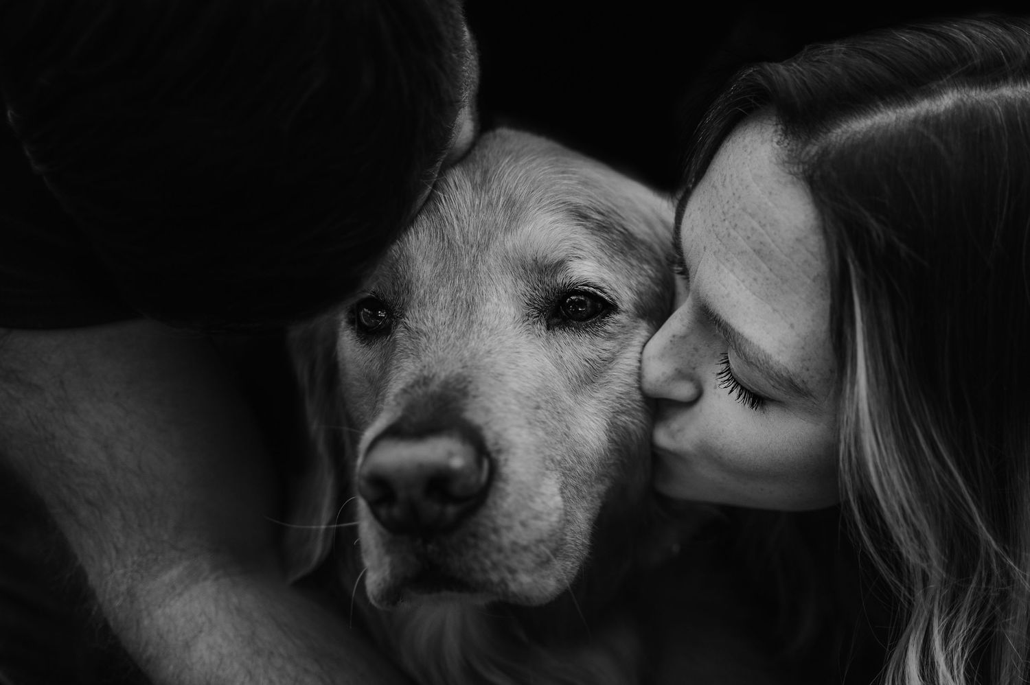Black and white photo of a couple kissing their golden retriever dog on the cheek in a tender moment.