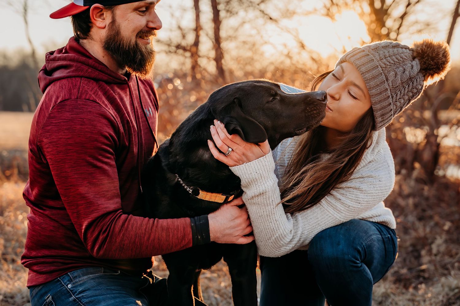 A couple in casual winter attire shares a warm moment with their black Labrador dog outdoors at golden hour.