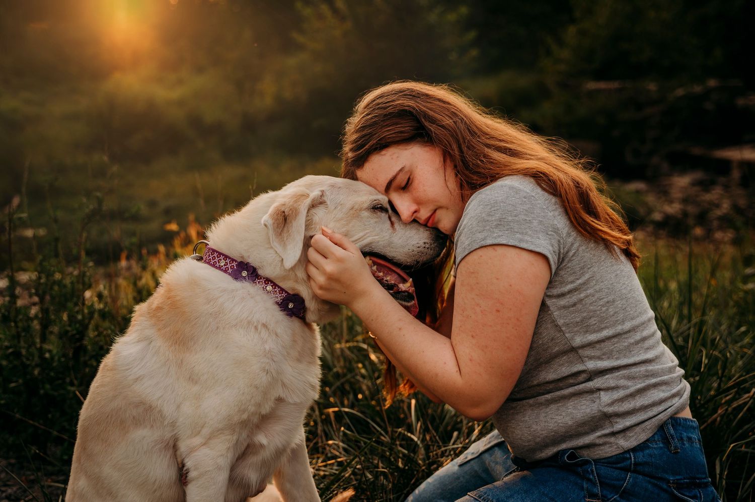 A young woman with auburn hair tenderly nuzzles a white Labrador dog outdoors in golden sunset light in Loudoun County Virginia.