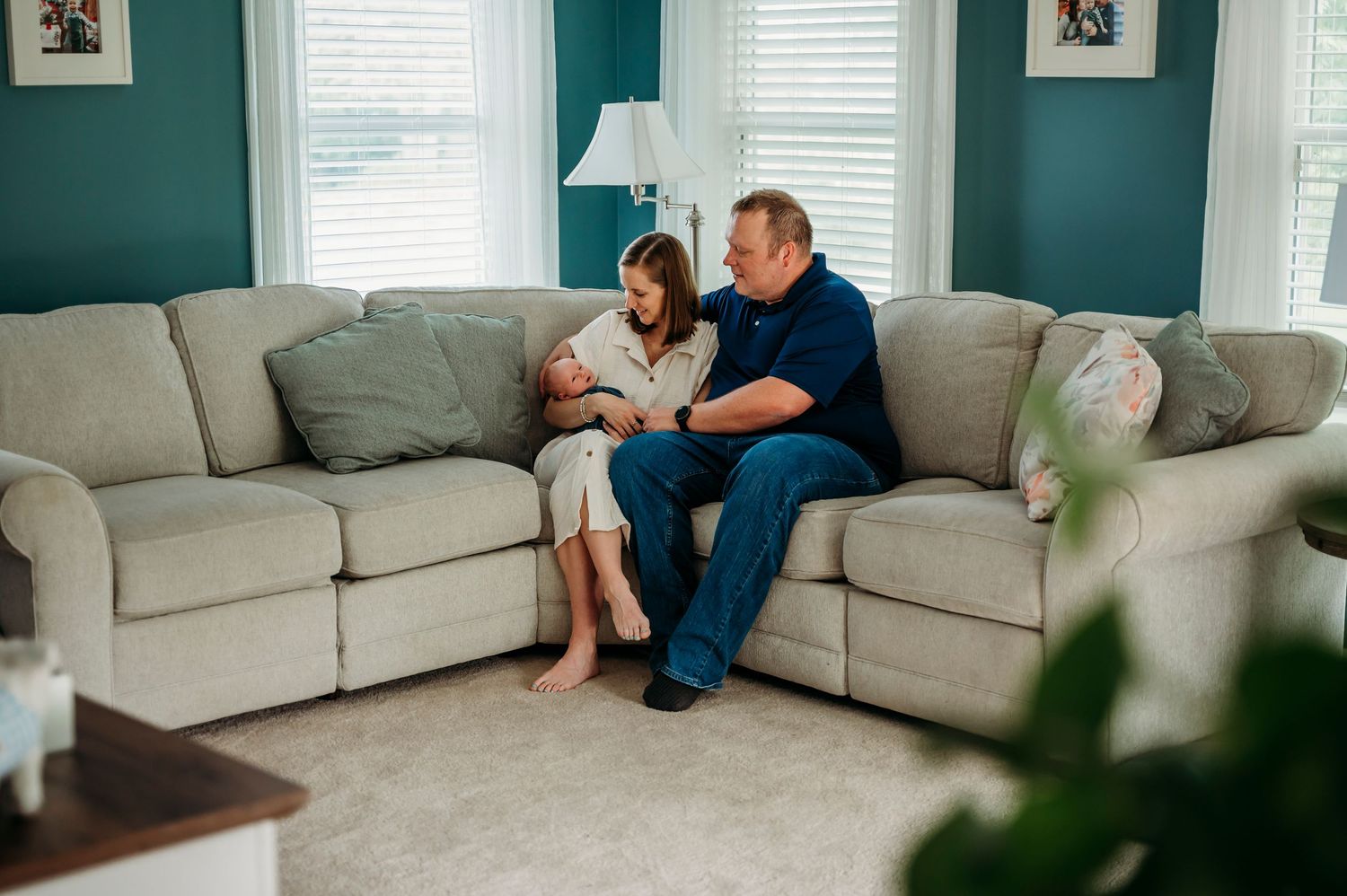 A couple sits together on a sofa in a living room during a lifestyle newborn session with their baby in Berryville Virginia.