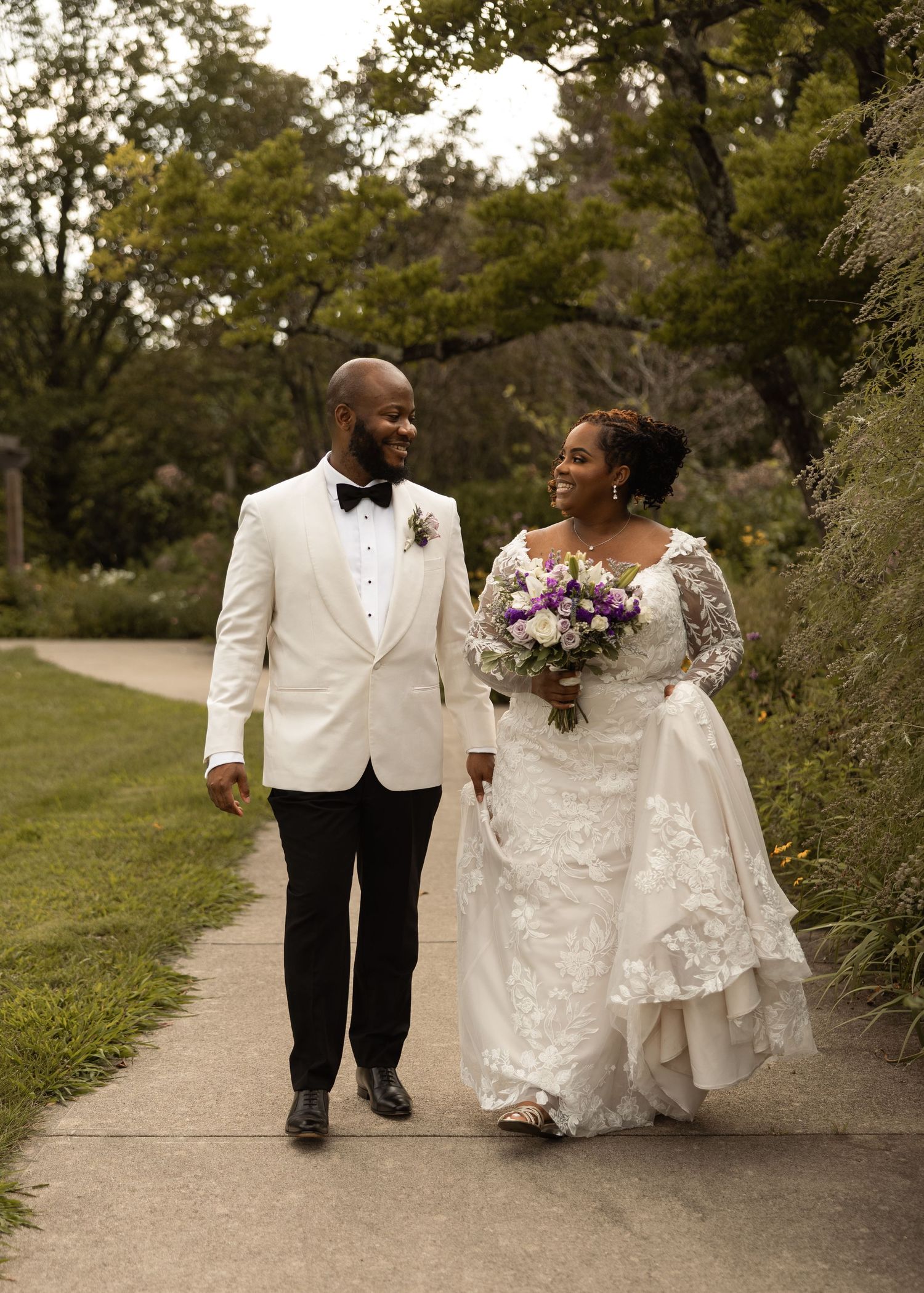 A smiling bride and groom walk hand-in-hand on a garden path, she holding a purple bouquet in wedding attire.