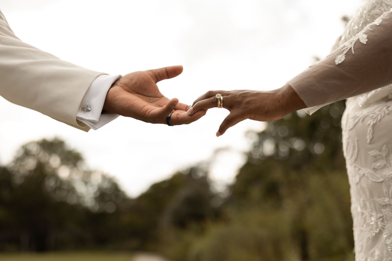Bride and groom reaching toward each other, wedding ring visible, outdoor ceremony backdrop.
