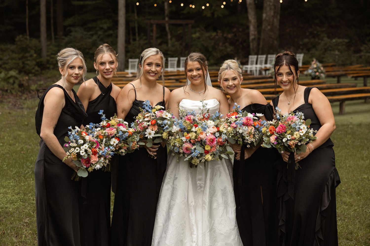 Bride in white gown with five bridesmaids in black dresses holding colorful floral bouquets outdoors at a wedding.