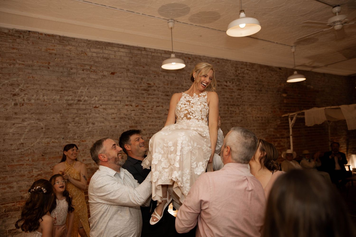 Bride lifted by guests during joyful wedding reception hora dance in rustic brick venue.