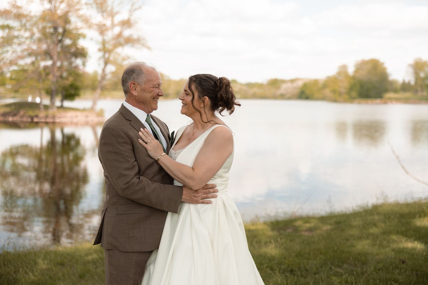 Bride and groom embrace near a serene lake, sharing a romantic moment in wedding attire outdoors.