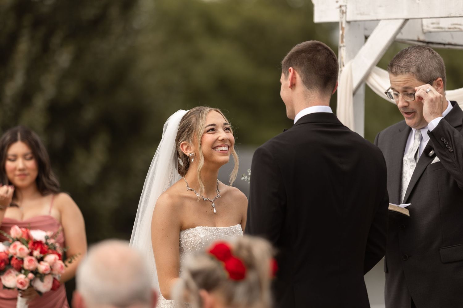Bride smiling joyfully at groom during outdoor wedding ceremony with bridesmaid holding red bouquet in background.