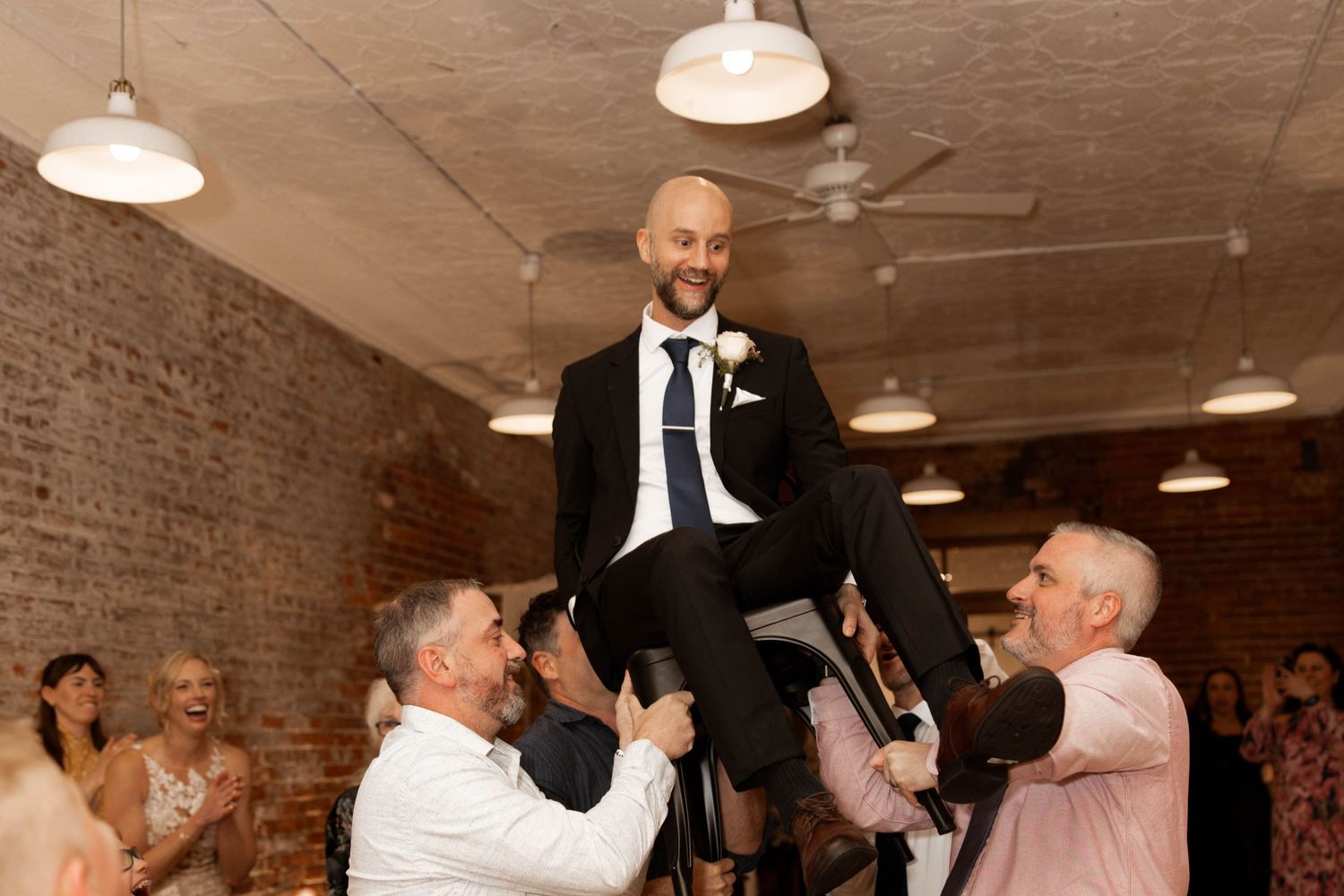 Groom in black suit lifted by groomsmen during wedding reception celebration in rustic brick venue.