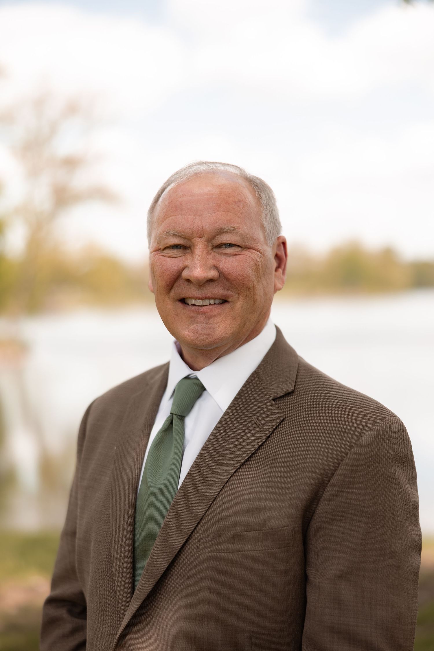 Professional headshot of a smiling middle-aged man in a brown suit and green tie, posed outdoors near a lake.