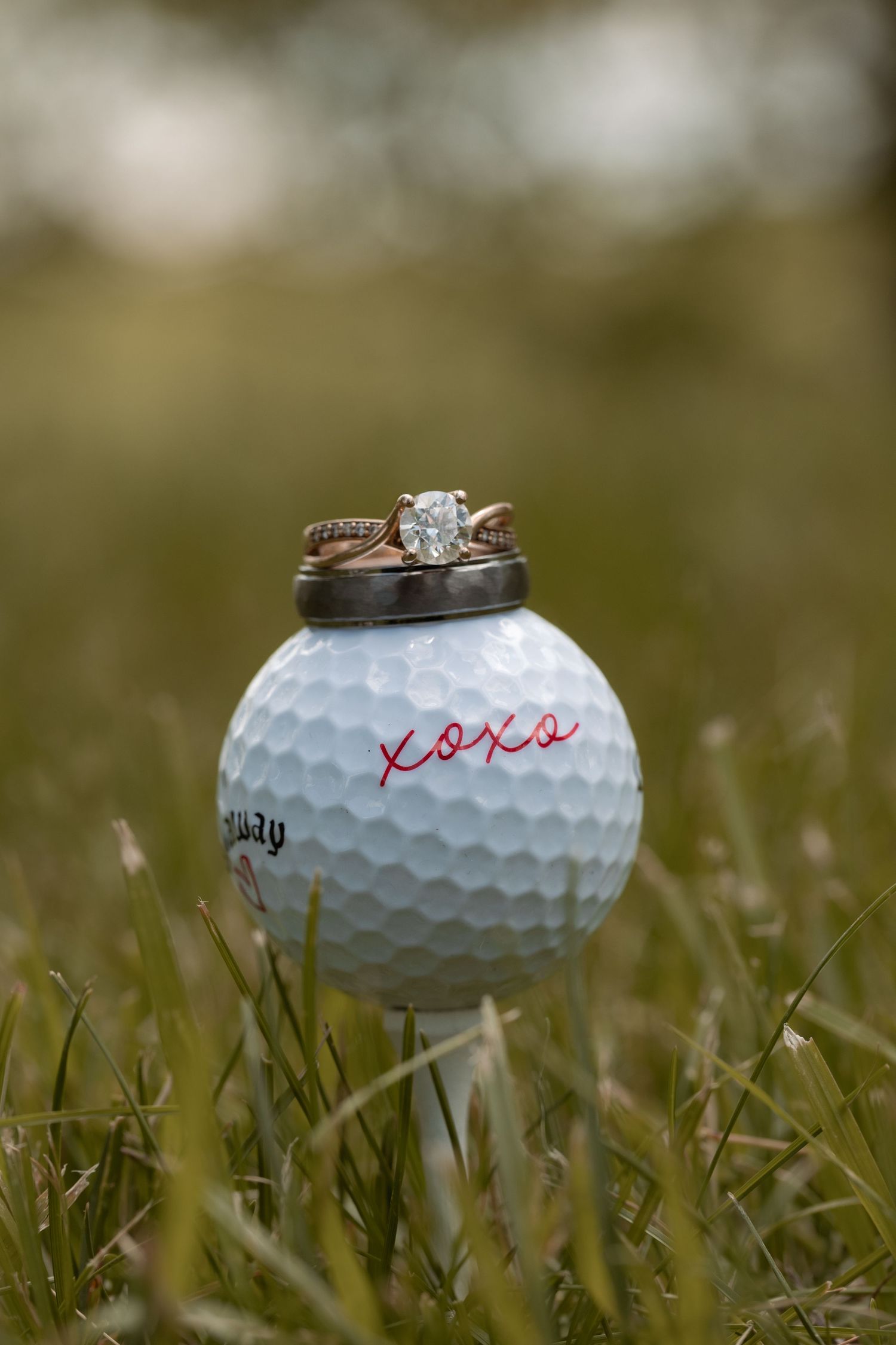 Wedding rings resting on a Callaway golf ball inscribed with XOXO, sitting in green grass.