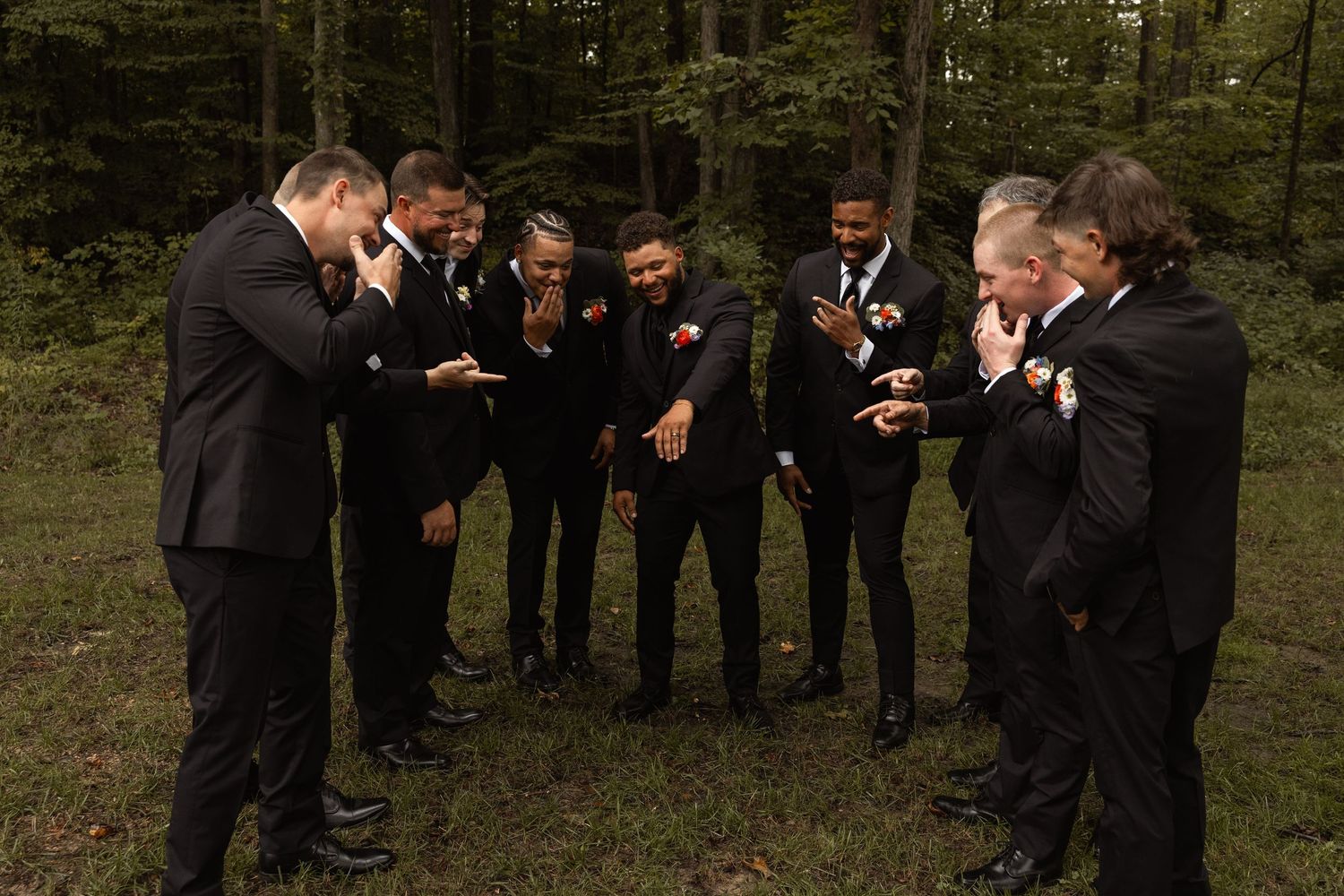 Groomsmen in black suits laughing together in a forest setting during a wedding celebration.
