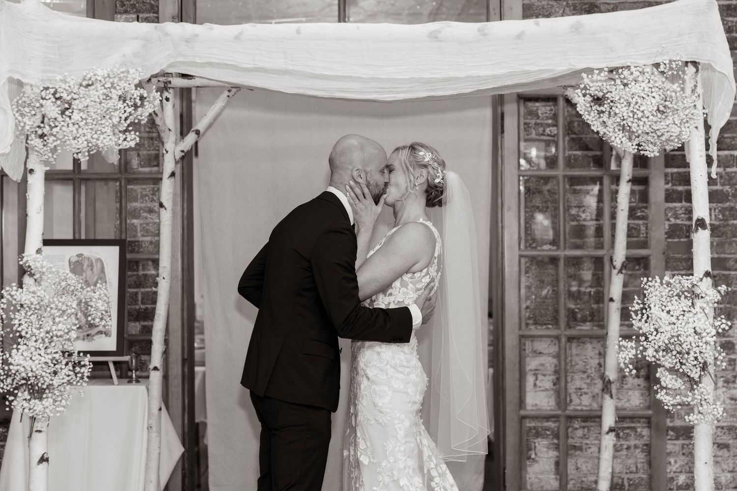 Black and white photo of a bride and groom sharing their first kiss under a floral wedding arch.