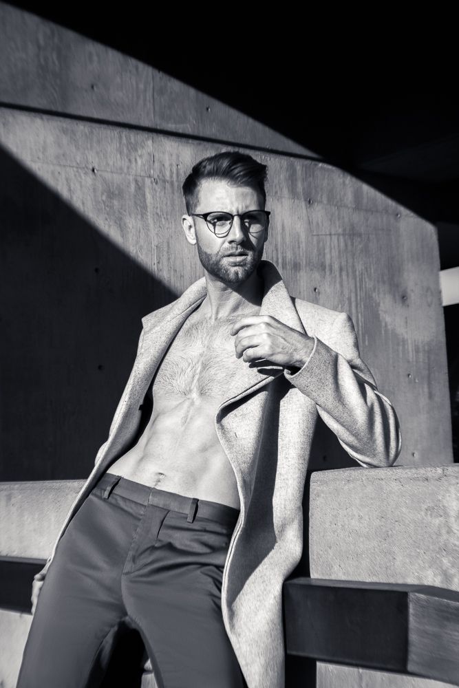 Muscular man in open coat and glasses posing shirtless in black and white outdoor photo.