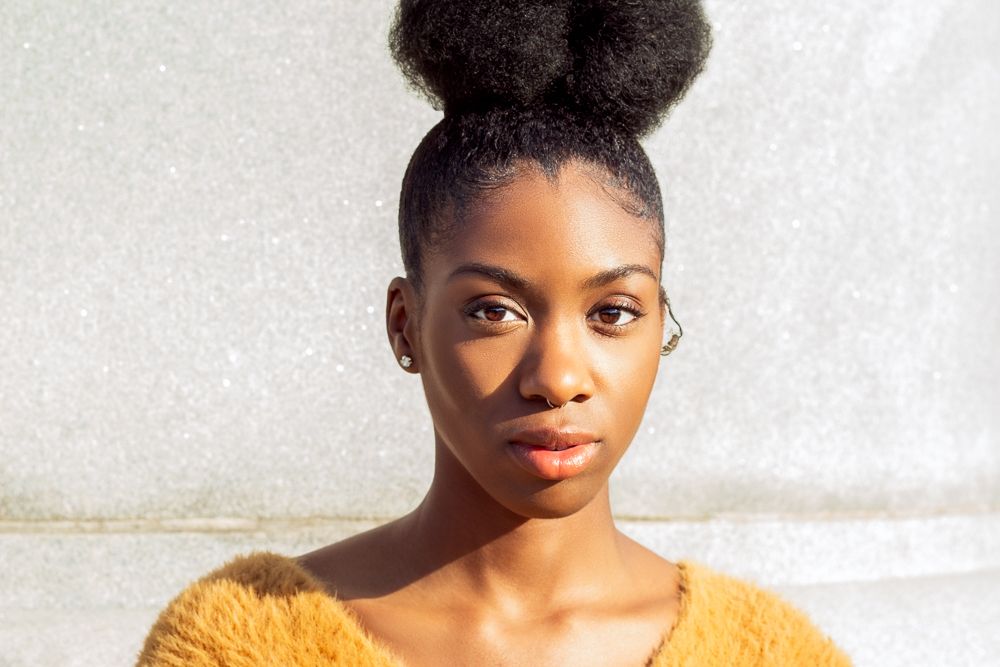 Young Black woman with natural hair bun in mustard fuzzy sweater against white wall.