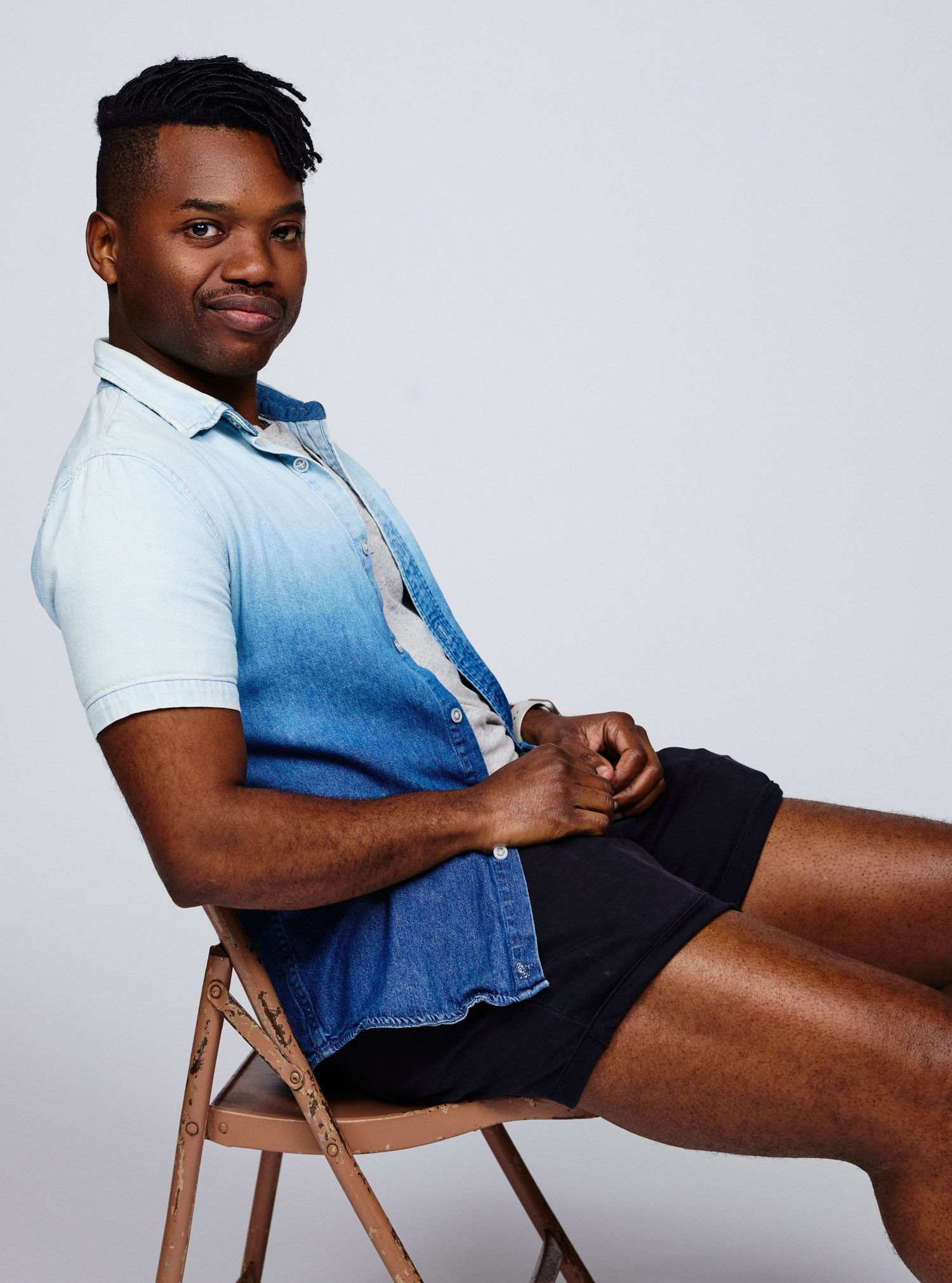Young man sitting on a folding chair wearing a blue ombre button-up shirt and black shorts, posing confidently.