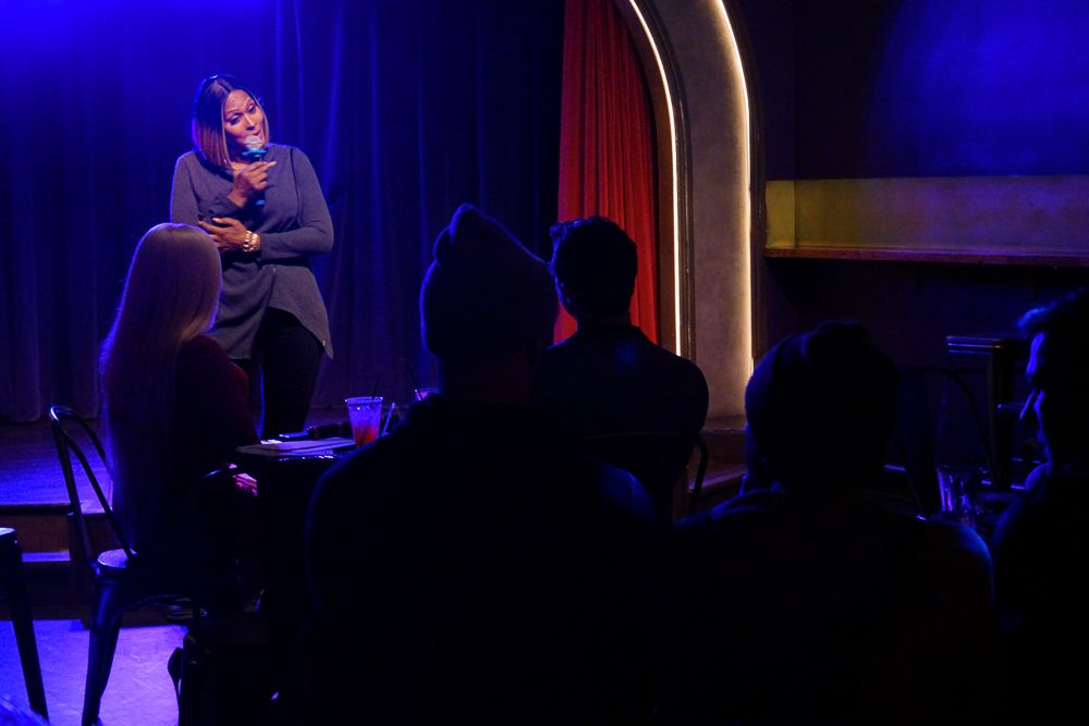 Performer on stage in dimly lit comedy club with red curtain backdrop and audience silhouettes in foreground.