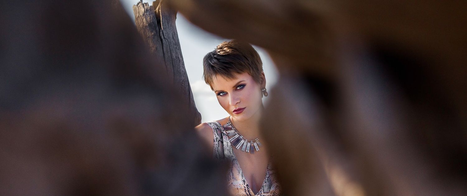 Young woman with short hair peers through blurred foreground elements in soft natural light.